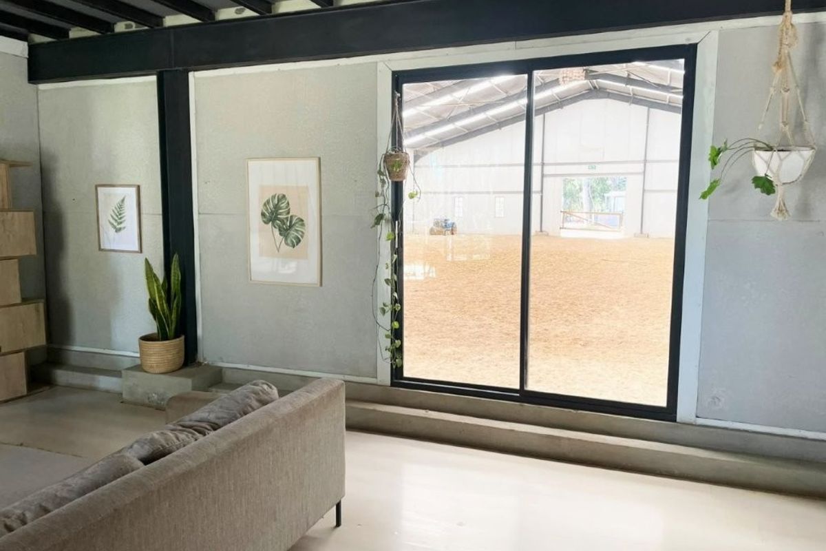 Modern living area featuring exposed steel structural beams, light timber flooring, and large sliding glass doors overlooking an indoor equestrian arena.