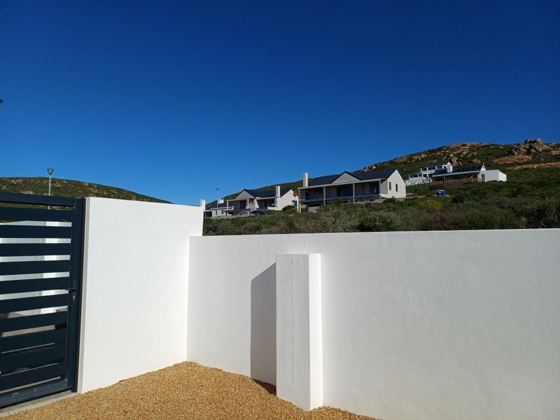 A view of the property entrance featuring a clean white boundary wall, a modern metal gate, and a gravel driveway area.