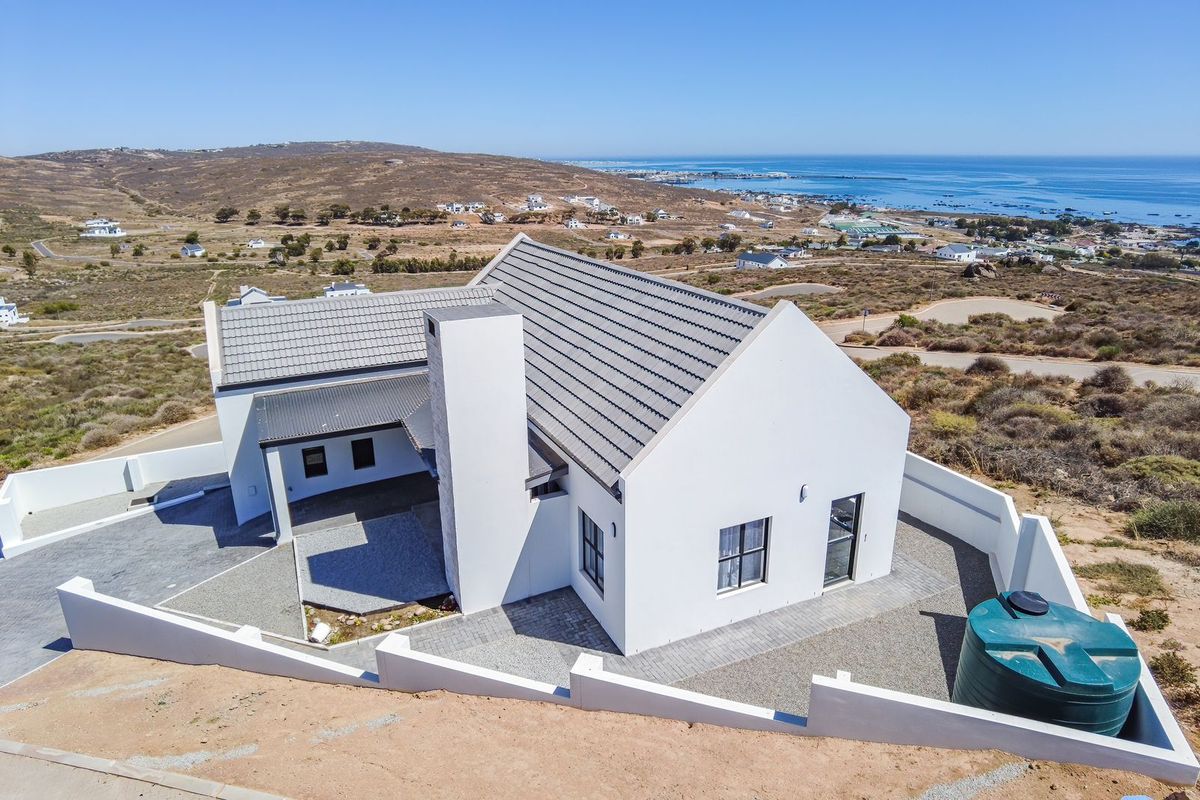 Aerial view of a modern coastal home featuring a tiled roof, prominent chimney, paved exterior areas, and a water tank within a walled perimeter.