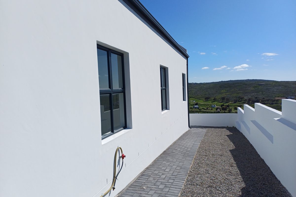 A clean exterior side view of the property featuring a paved walkway, gravel landscaping, and a modern white facade with dark aluminium window frames.