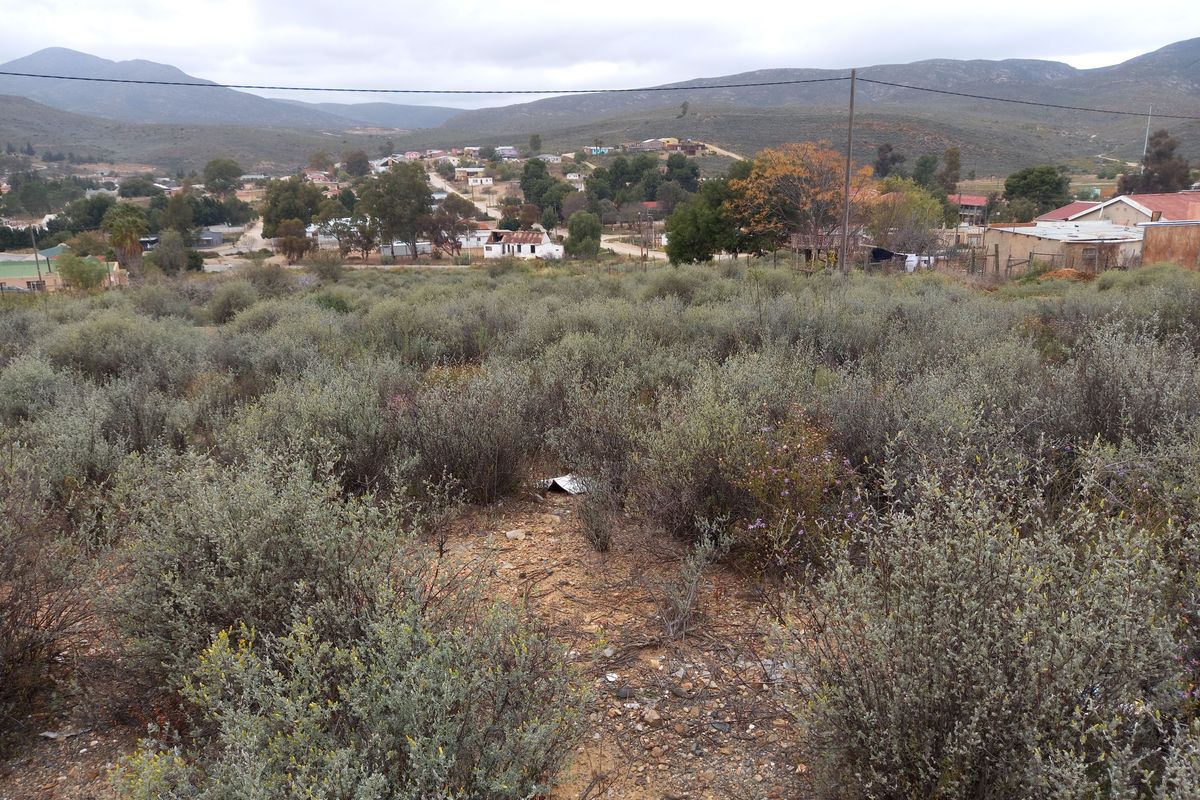 A view overlooking a vacant residential plot with natural vegetation, featuring surrounding hills and distant neighborhood structures.