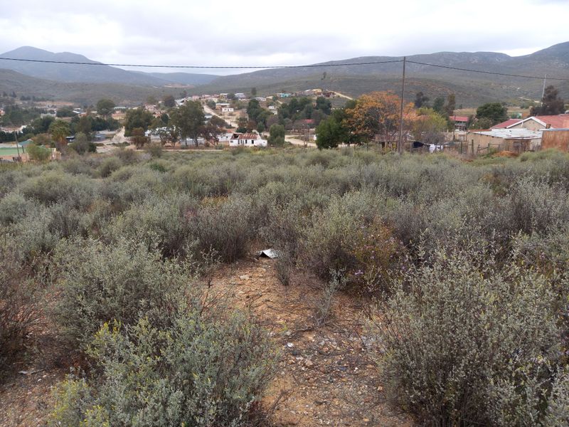 A view overlooking a vacant residential plot with natural vegetation, featuring surrounding hills and distant neighborhood structures.