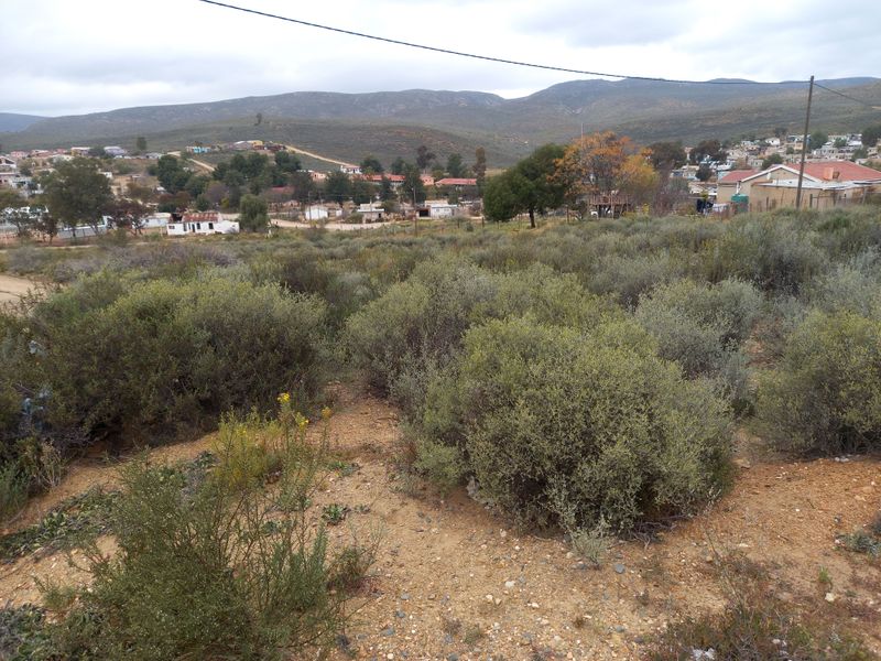 A view of a vacant residential plot featuring natural vegetation and sloped terrain with surrounding neighborhood and mountain views.