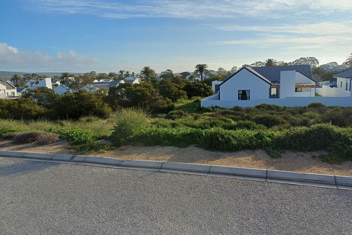 A street-level view of a vacant residential plot featuring native vegetation, with established neighboring homes visible in the background.