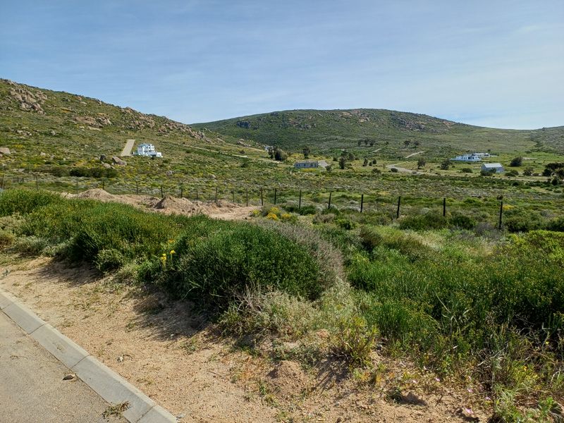 A wide-angle view of a vacant residential plot featuring natural fynbos vegetation, rolling hills, and distant neighboring properties.