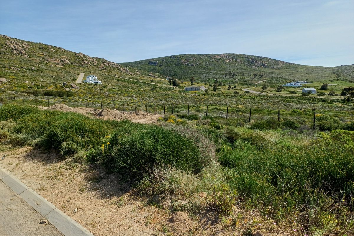 A wide-angle view of a vacant residential plot featuring natural fynbos vegetation, rolling hills, and distant neighboring properties.