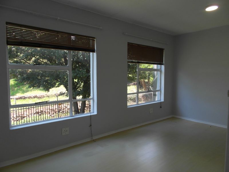A bright, empty living room featuring light-toned laminate flooring, neutral walls, and large windows with fitted venetian blinds.