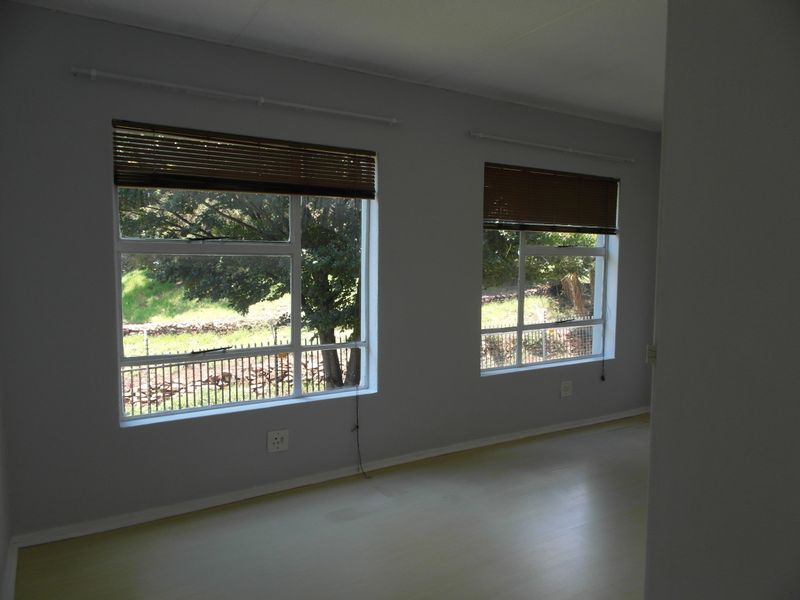 An empty bedroom featuring laminate flooring, neutral grey walls, and two windows with fitted venetian blinds overlooking the exterior grounds.