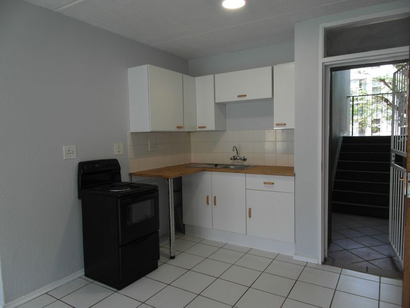 A compact, functional kitchen featuring white cabinetry, a laminate countertop, tiled backsplash, and ceramic floor tiles, with direct access to an exterior stairwell.