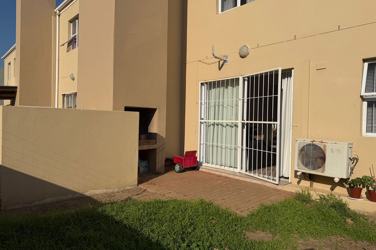 Rear exterior view of the townhouse featuring a paved patio area, built-in braai, and access to the interior through security-gated sliding doors.