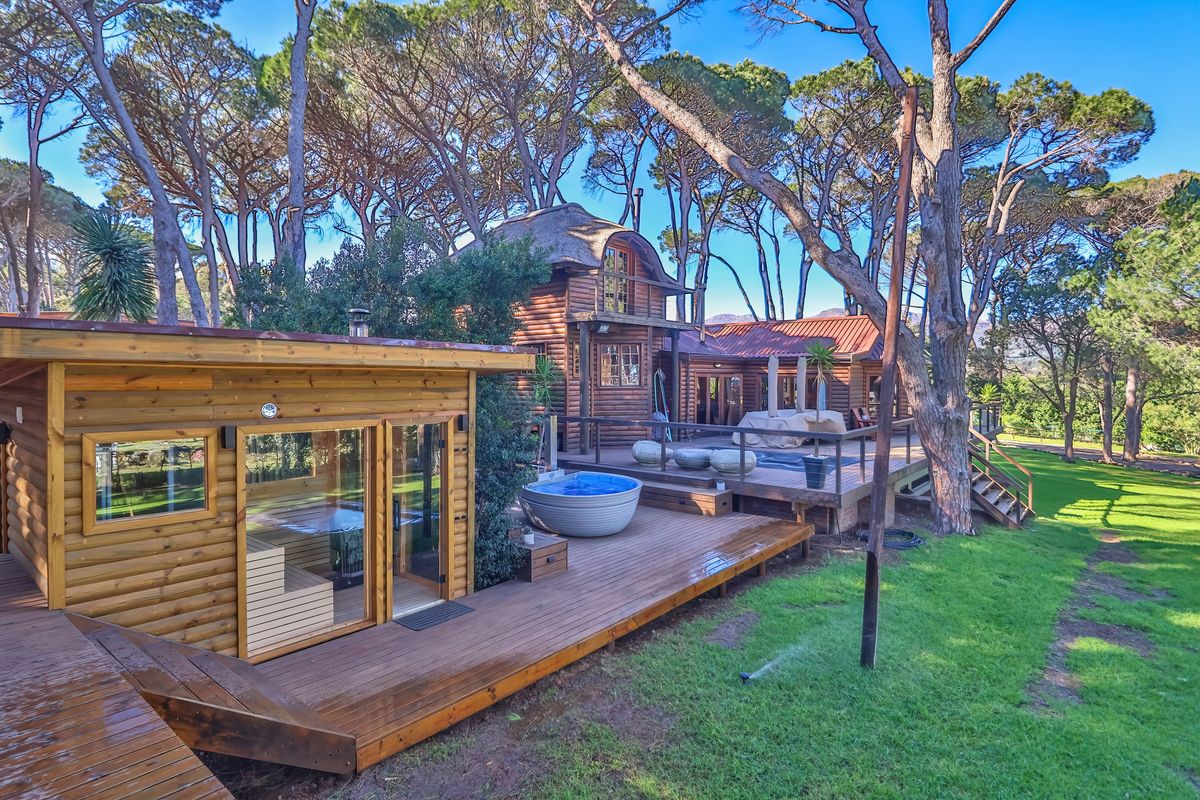 A scenic rear exterior view featuring a timber deck, a standalone sauna, a hot tub, and a rustic log cabin dwelling set among mature trees.