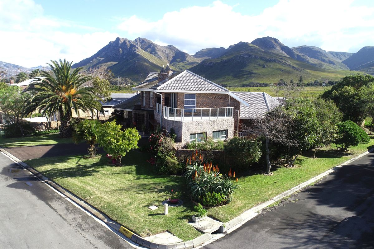 Aerial view of a double-story brick house featuring a balcony, tiled roof, and landscaped garden, set against a scenic mountain backdrop.