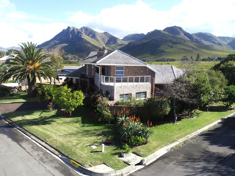 Aerial view of a double-story brick house featuring a balcony, tiled roof, and landscaped garden, set against a scenic mountain backdrop.