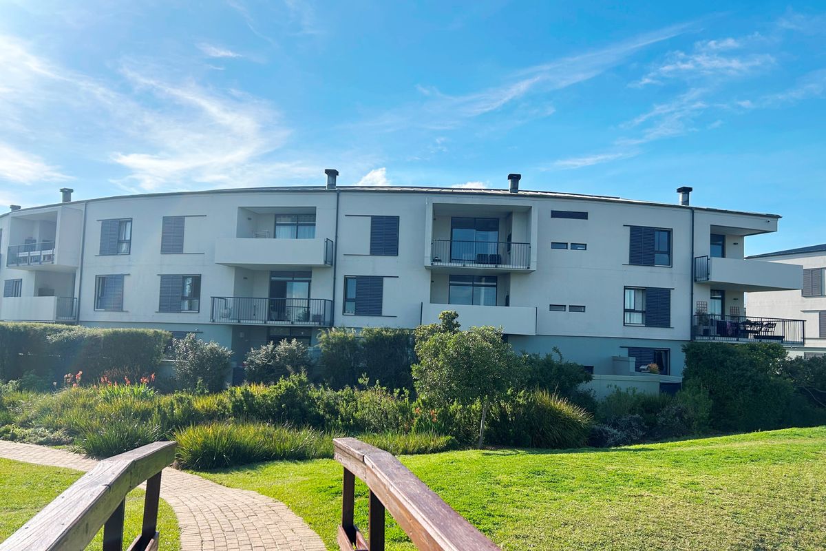 Rear view of a modern apartment complex featuring multiple balconies, sliding glass doors, and a landscaped garden area with a pedestrian bridge.