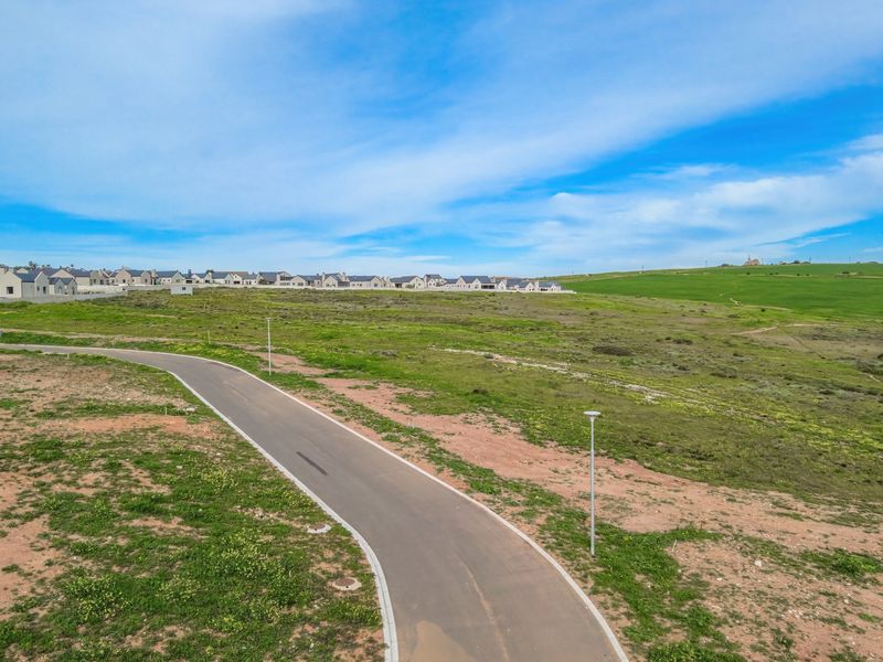 An aerial view of a vacant residential plot featuring a paved access road and street lighting, situated within a developing estate.