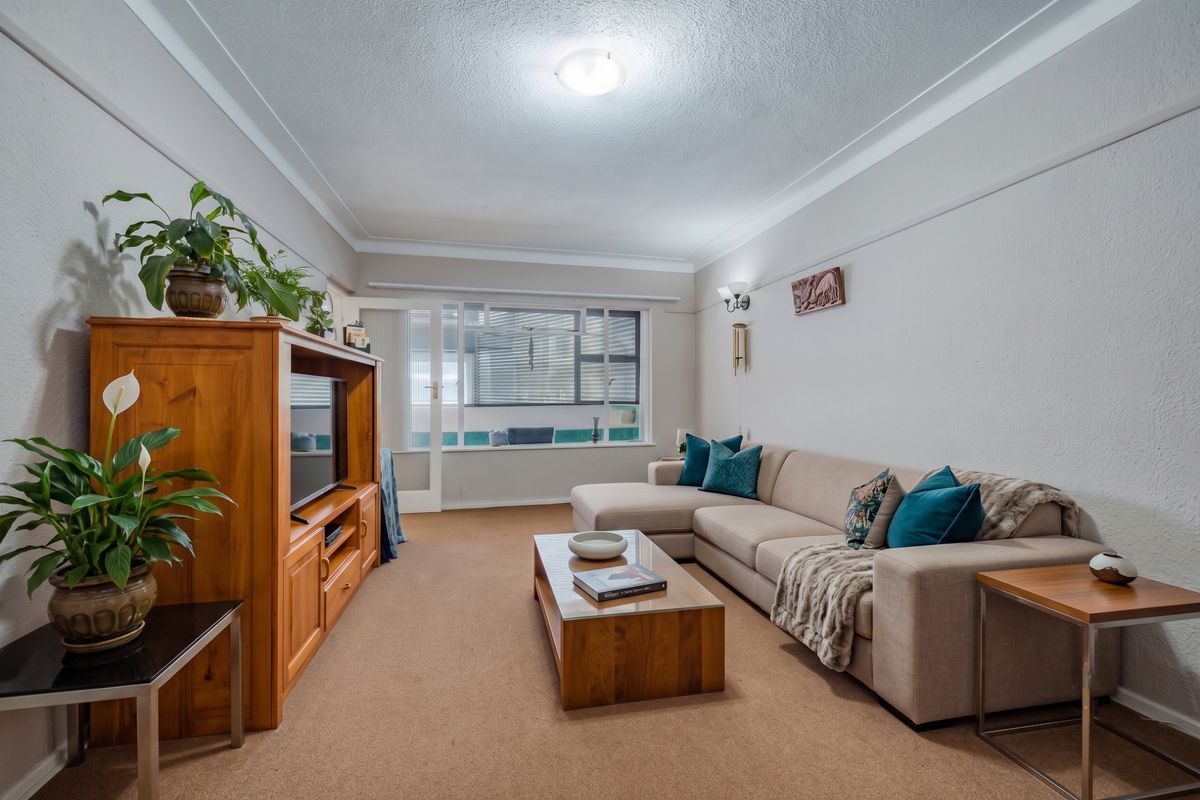 A comfortable living room featuring neutral walls, carpeted flooring, and a large window allowing natural light, with access to an adjacent balcony area.