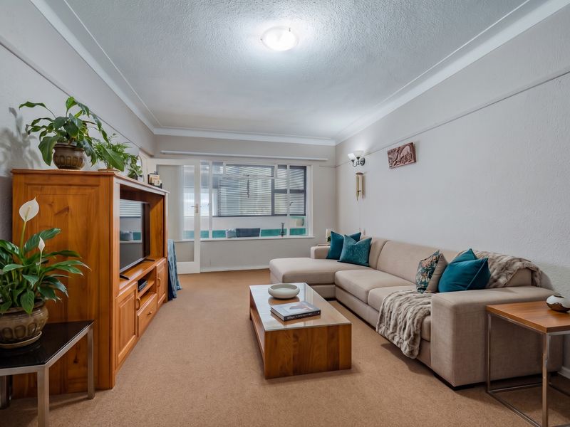 A comfortable living room featuring neutral walls, carpeted flooring, and a large window allowing natural light, with access to an adjacent balcony area.