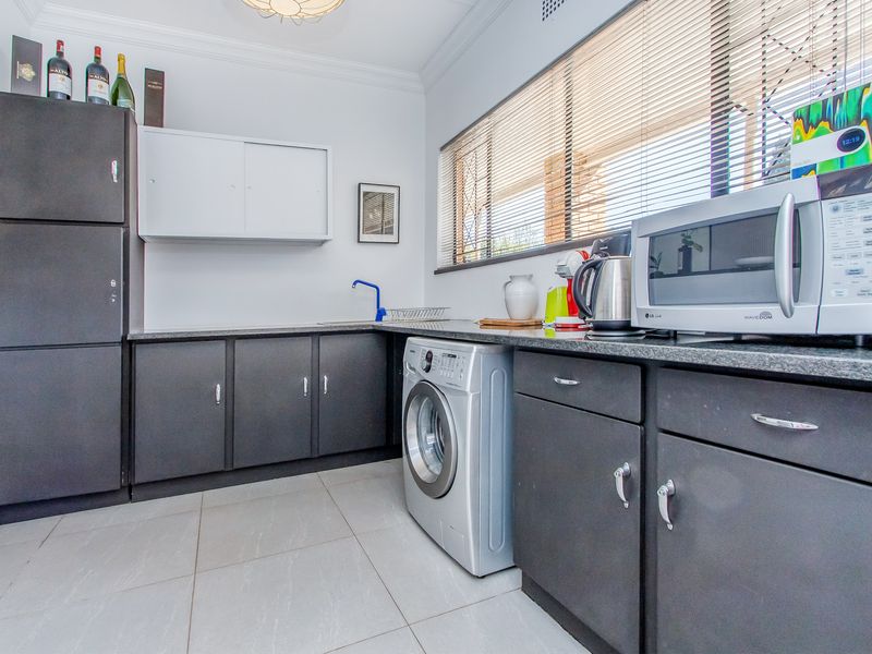 A functional laundry room featuring dark built-in cabinetry, granite countertops, a stainless steel sink, and tiled flooring with large windows and venetian blinds.