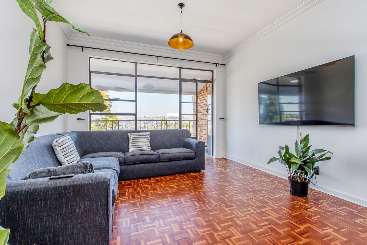 Bright living room featuring classic parquet flooring, large steel-framed windows providing natural light, and access to an outdoor balcony area.