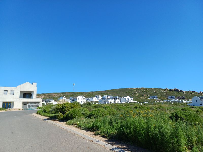 A street-level view of a residential area featuring an asphalt road, curb infrastructure, and surrounding coastal landscape with nearby homes.