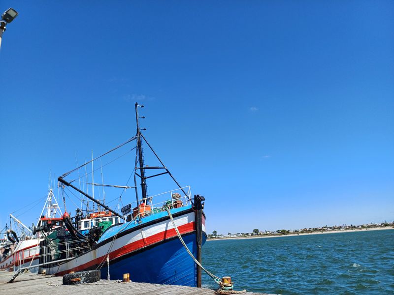 A view of a fishing boat docked at a pier with a coastal landscape in the background.