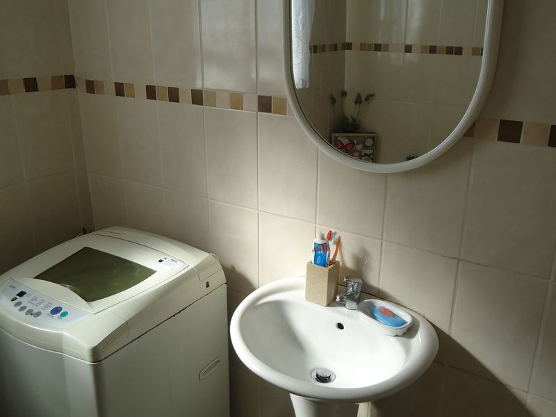 A functional laundry area featuring a pedestal basin, wall-mounted mirror, and neutral ceramic wall tiling.