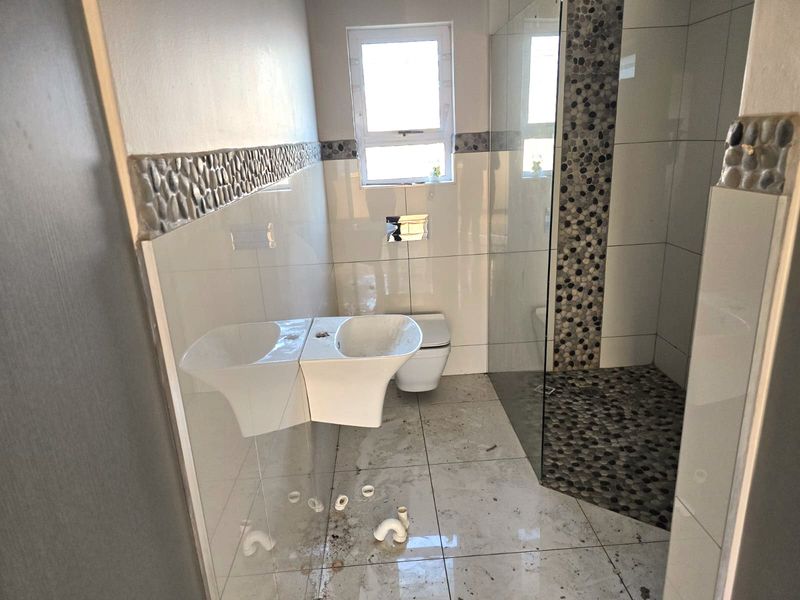 A bathroom in need of completion, featuring a glass shower enclosure, wall-mounted toilet, and pebble tile accents, currently missing a vanity basin.