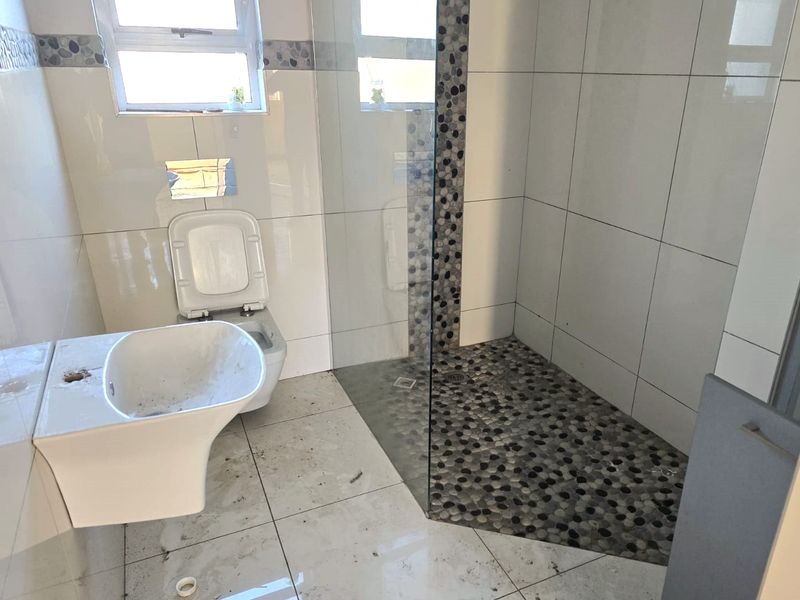 A bathroom featuring a wall-mounted toilet, glass shower enclosure with pebble flooring, and tiled walls. The space requires completion and cleaning.