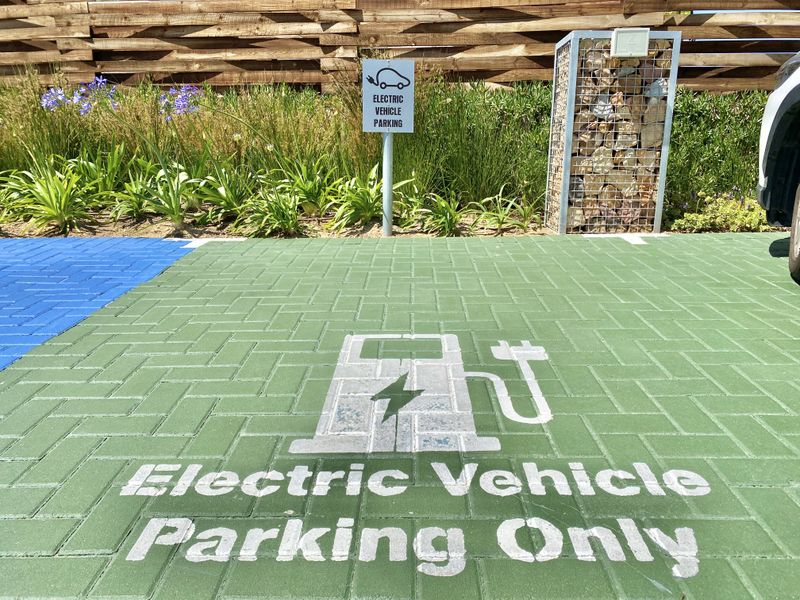 A designated electric vehicle parking bay featuring green painted paving, clear signage, and a modern gabion wall with timber fencing in the background.