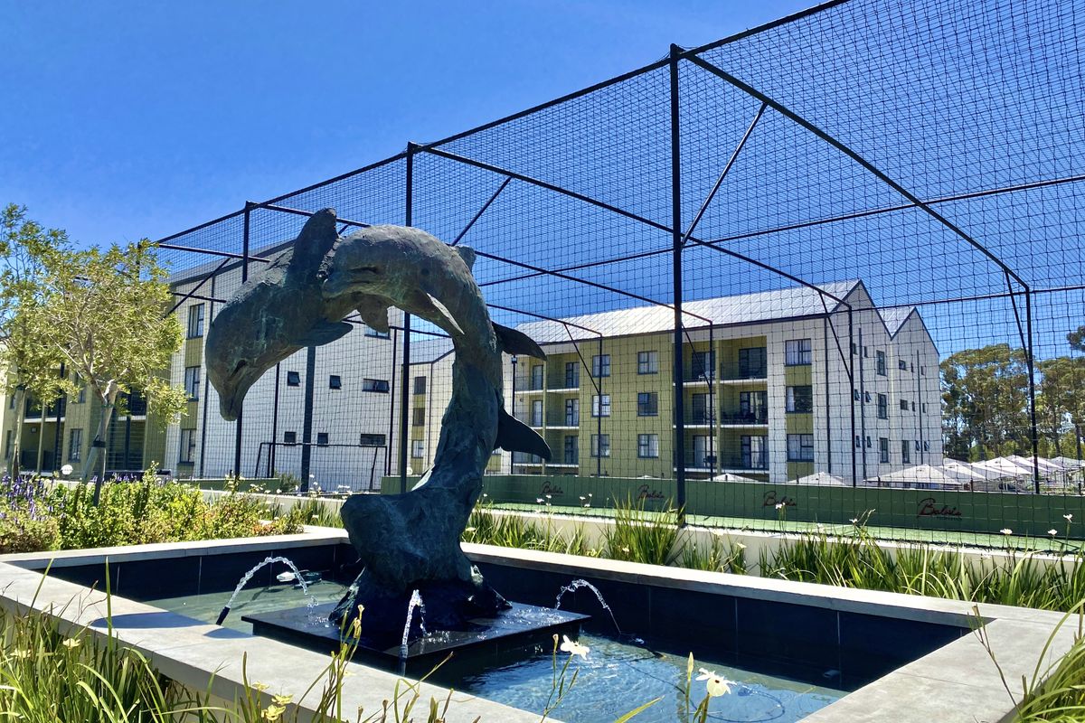 A well-maintained outdoor area featuring a decorative dolphin fountain, landscaped garden beds, and sports netting adjacent to the apartment building complex.