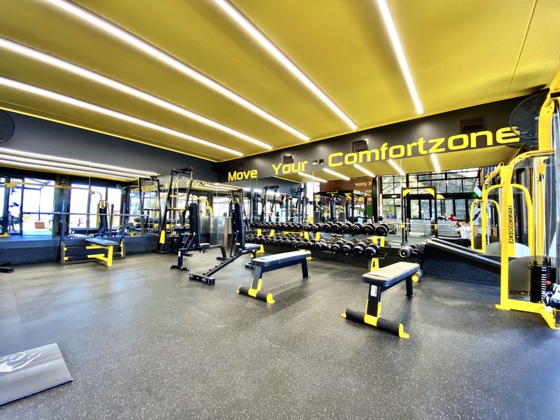 A modern, well-equipped gym facility featuring professional rubberized flooring, extensive mirrored walls, and bright, recessed linear ceiling lighting.