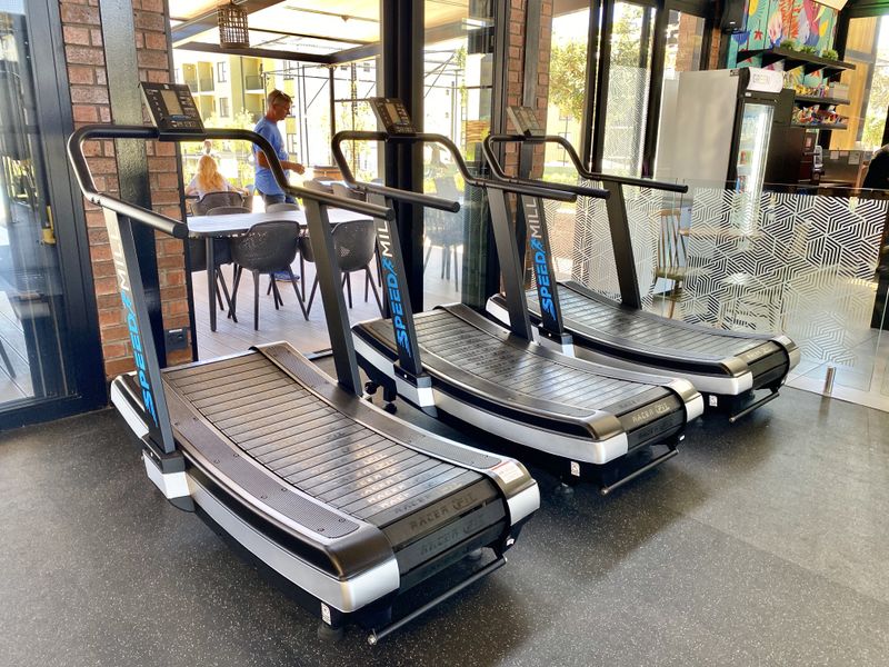 A modern gym facility featuring rubber flooring, exposed brick accents, and large glass sliding doors opening to an outdoor patio area.