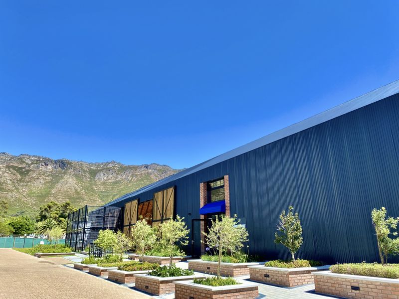 Modern industrial-style building exterior featuring corrugated metal cladding, brick planter boxes, and a paved walkway against a scenic mountain backdrop.