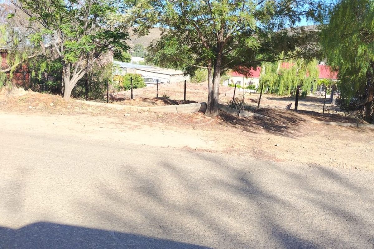 A street-level view of a vacant residential plot featuring mature trees and perimeter wire fencing along an asphalt road.