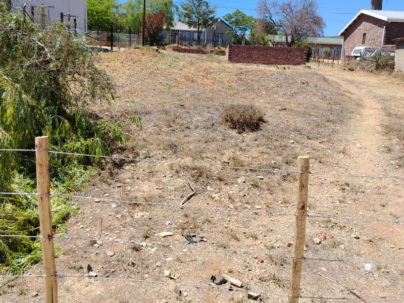 A view of a vacant residential plot featuring a wire fence, wooden posts, and a brick retaining wall in a suburban setting.