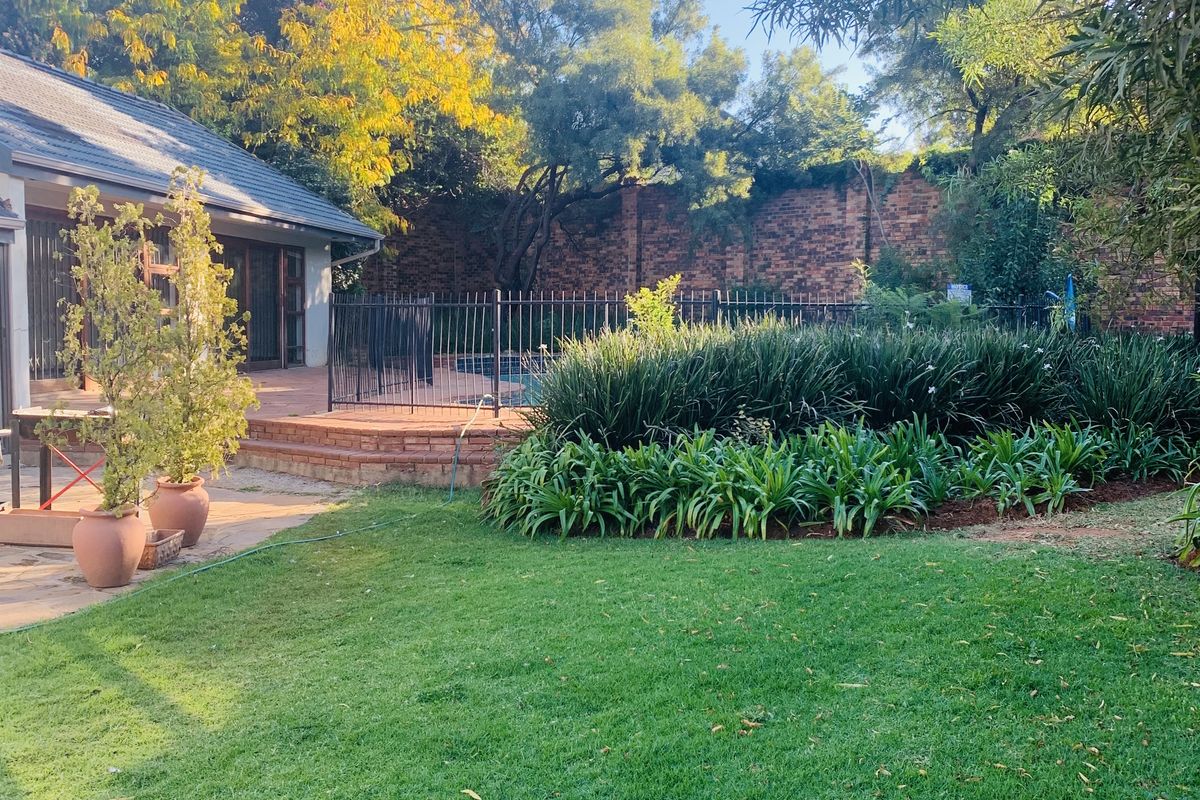 A well-maintained rear garden featuring a lush lawn, landscaped garden beds, a paved patio area, and a fenced swimming pool.