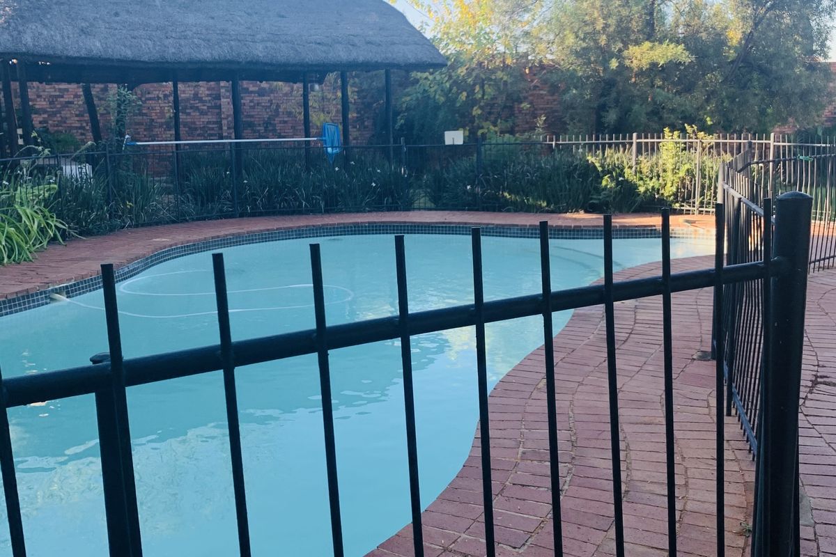 Outdoor pool area featuring a swimming pool, brick paving, and a thatched lapa structure, enclosed by metal safety fencing.
