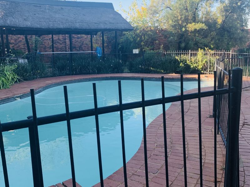 Outdoor pool area featuring a swimming pool, brick paving, and a thatched lapa structure, enclosed by metal safety fencing.