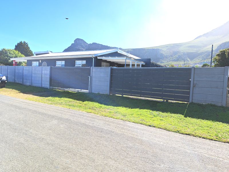 Front exterior view of a single-story house featuring a modern perimeter fence, sliding gate, and corrugated metal roof set against a mountain backdrop.