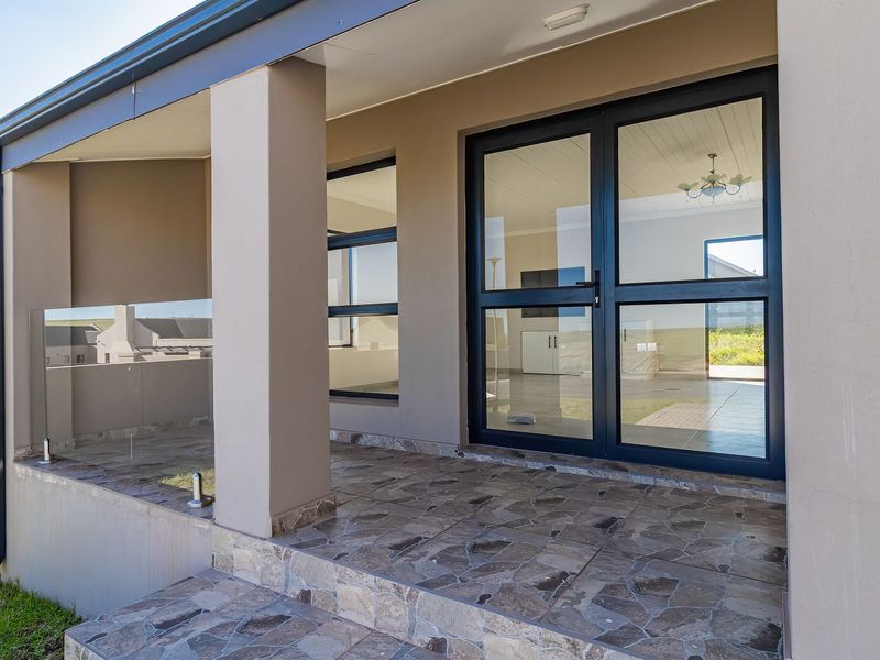 A modern covered patio featuring stone tile flooring, a sleek glass balustrade, and double glass doors leading into the interior living space.