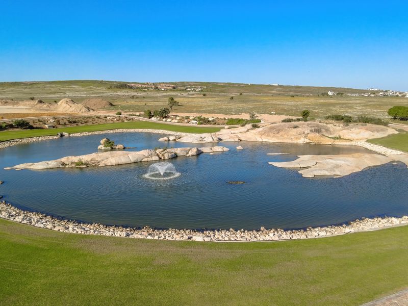 An elevated aerial view of a landscaped pond with a central water fountain, rock features, and surrounding manicured golf course grounds.