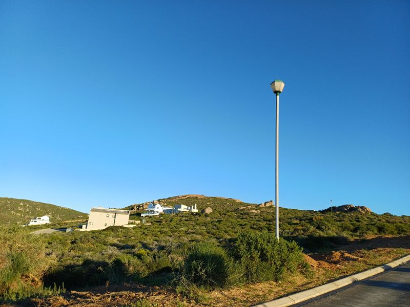 A view of a vacant residential plot featuring natural vegetation, street lighting, and paved road infrastructure with surrounding hillside landscape.