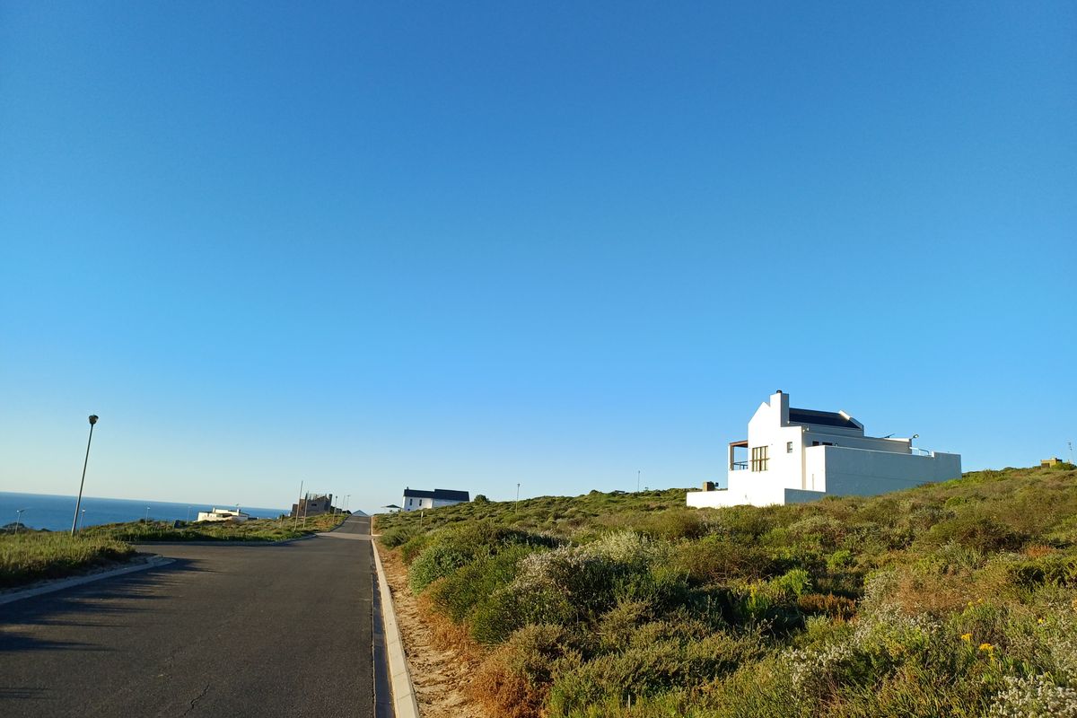 A view of the paved residential street and surrounding coastal landscape with a modern white house visible in the distance.