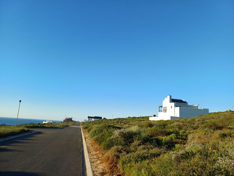 A view of the paved residential street and surrounding coastal landscape with a modern white house visible in the distance.
