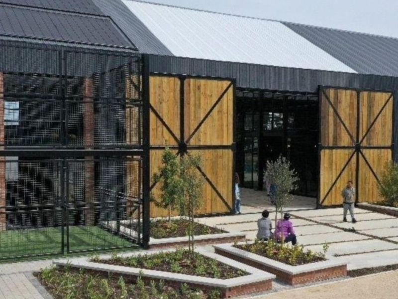 Modern industrial-style building exterior featuring large sliding timber barn doors, metal roofing, brick accents, and landscaped concrete paved entrance area.