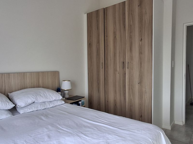 A well-maintained bedroom featuring a built-in wooden wardrobe, neutral wall finishes, and tiled flooring, leading into a hallway entrance.