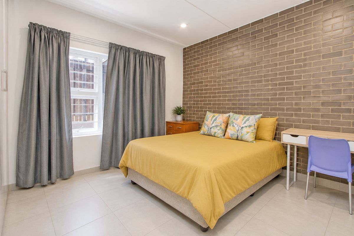 A well-maintained bedroom featuring a distinctive exposed brick feature wall, neutral tiled flooring, and a window fitted with security bars.