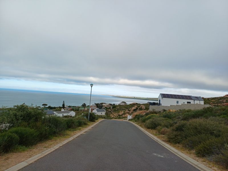 A view looking down a paved residential street towards the coastline, featuring nearby homes and clear ocean views.
