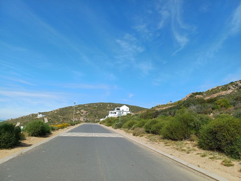A view of the paved access road leading through the residential estate, surrounded by natural coastal vegetation and hilly terrain.