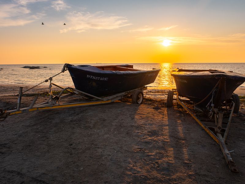 A scenic sunset view over the ocean featuring boats on trailers parked on the sandy shoreline.